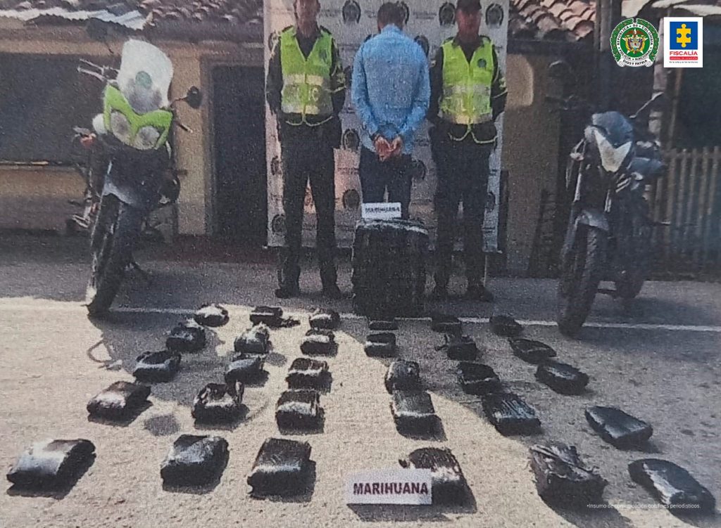 En la foto aparece de pie y de espaldas a la cámara Roberth Smith Márquez Ríos. Esta persona viste camisa de manga larga azul claro y pantalón oscuro. A lado y lado de él hay uniformados de la Policía Nacional y dos motocicletas. En el piso hay varios paquetes negros y junto a ellos hay dos letreros que dicen: ‘Marihuana’. Igualmente roja de manga corta y pantalón oscuro. En la parte posterior hay un pendón institucional de la Policía.