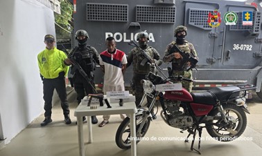 Se visualiza al capturado junto a uniformados de la Policía y Ejército Nacional. Frente a ellos los elementos incautados como armas, granda, proveedor, celular y una motocicleta.
