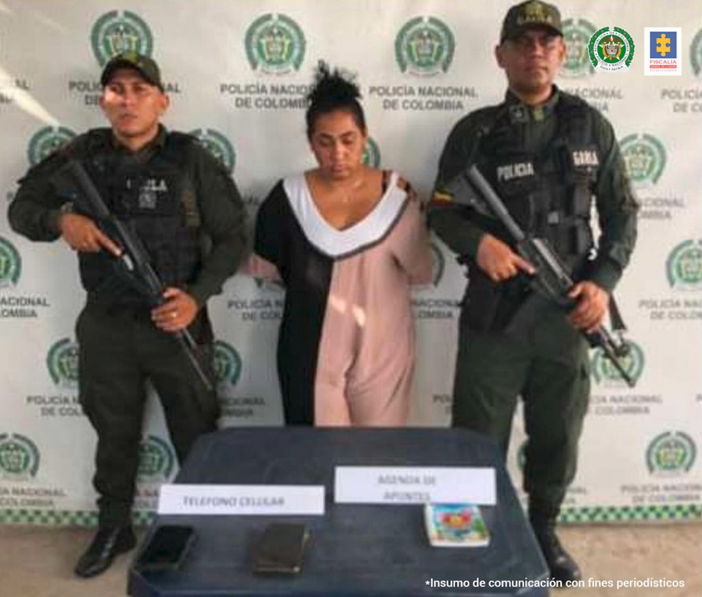 En la fotografía aparece una mujer capturada, acompañada de dos uniformados de la Policía Nacional. En la parte posterior un banner con logos de la entidad.