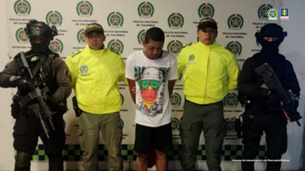 En la fotografía se aprecia de pie y con los brazos atrás a Rolber Quiñones Araujo, alias Lágrima. Viste camiseta de color blanco, con esampado grande de colores; bermuda de color negro. En sus extremos hay uniformados de la Policía Nacional. Detrás de ellos hay un pendón institucional de la Policía.