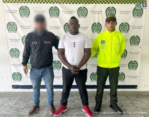 En la fotografía se aprecia de pie, con los brazos adelante y esposado a Ronald Pereira Landázuri. Viste camiseta blanca con estampado a la altura con las palabras ‘Tommy Hilfiger’, separadas del logo de la misma; pantalón negro y zapatillas rojas con zuela blanca. Al lado derecho de él hay un servidor del CTI de la Fiscalía y a su izquierda un uniformado de la Policía Nacional. Detrás de ellos hay un pendón institucional de la Policía.