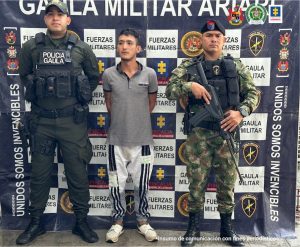 capturado de frente manos atrás esposado custodiado por un policía y un milkitar. Detrás banner de Gaula Militar de Ejército y Fiscalía.
