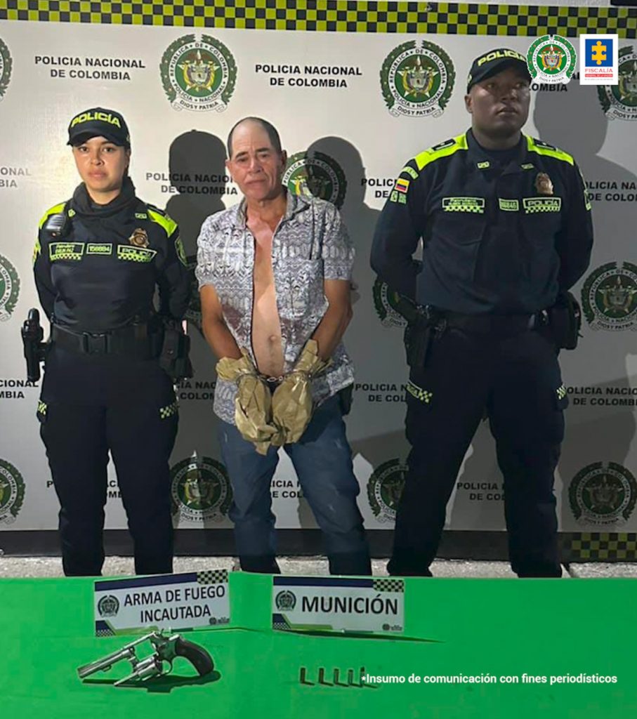 En la foto se aprecia de pie, con los brazos adelante, esposado y sus manos envueltas en bolsas a Rubén De Jesús Cruz Ocampo. Viste camisa desabotonada, de manga corta estampada con negro y blanco; jean azul. A lado y lado de él hay uniformados de la Policía Nacional. Delante de ellos hay una superficie verde y sobre ella se aprecia un arma de fuego y munición. Asimismo, dos letreros que dicen: ‘Arma de fuego incautada’, y ‘Munición’. Detrás de ellos hay un pendón institucional de la Policía.