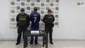 en la imagen se ve una persona detenida de espaldas bajo custodia de dos integrantes de la Policía Nacional. Detrás suyo un backing institucional.