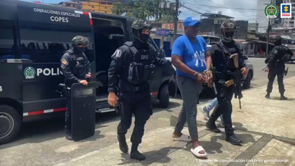 Hombre robusto de camiseta y gorra azul camina esposado. Es acompañado a lado y lado por dos uniformados de la Policía Nacional que lo conducen.
