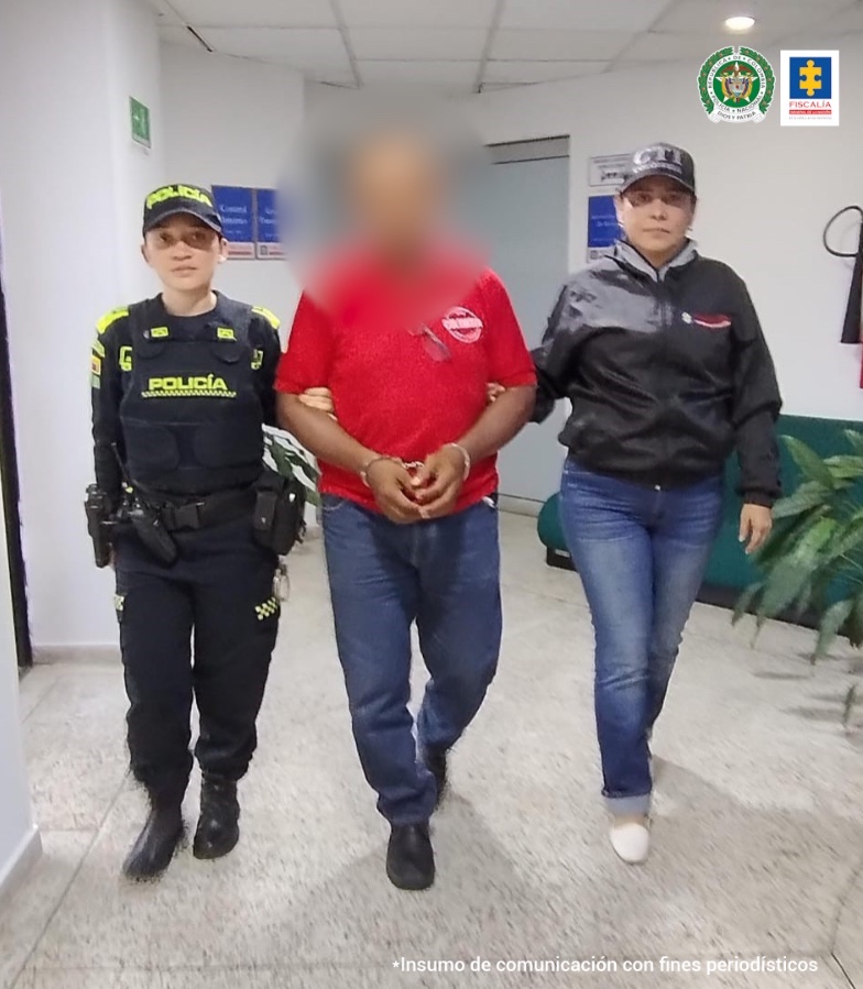 En la fotografía se aprecia en acción de caminata, con los brazos adelante y esposado a Sair Marino Mosquera Pérez. Viste camisa roja de manga corta, jean azul y zapatos negros. Al lado izquierdo de él hay una servidora del CTI de la Fiscalía y al derecho de él hay una uniformada de la Policía Nacional.