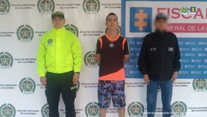 En la imagen se ve al capturado en custodia por un servidor del Cuerpo Técnico de Investigación CTI y un uniformado de la Policía Nacional. El investigado tiene un buzo negro con franjas negras y una bermuda estilo militar gris con azul.