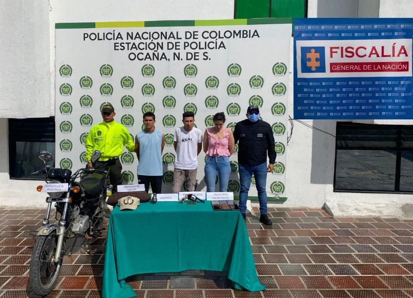 Se visualiza a los capturados junto a un uniformado de la Policía Nacional e investigador del CTI. Frente a ellos una mesa con los elementos incautados (un revólver, granada de fragmentación, teléfonos celulares y motocicleta). Detrás el banner que identifica a la Policía Nacional.