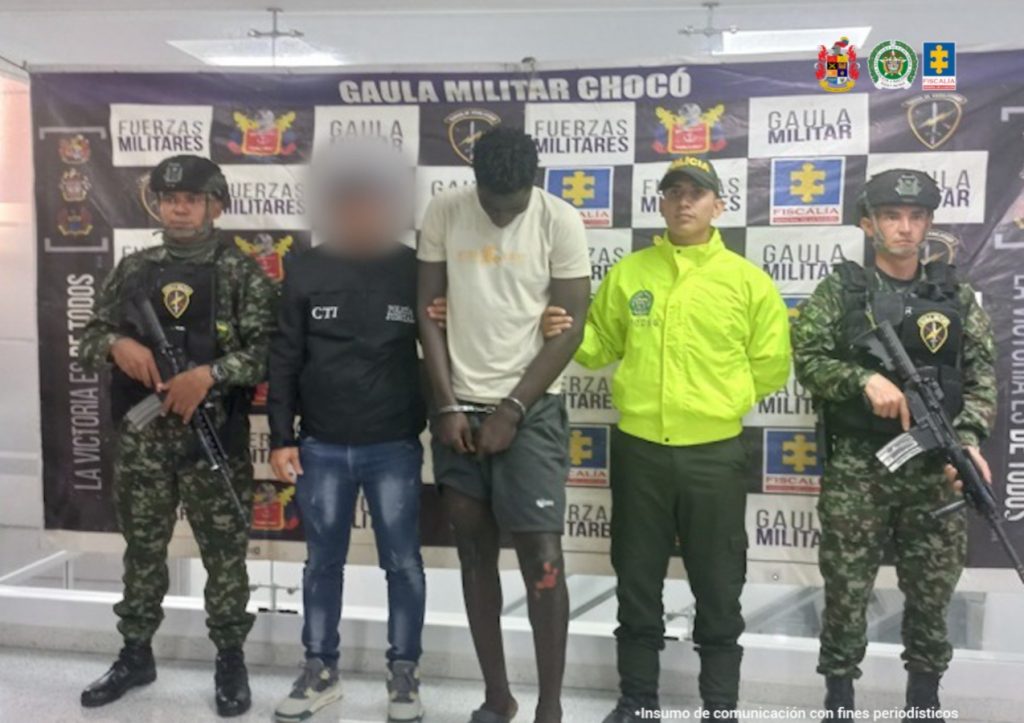 El capturado viste camiseta blanca y pantalón corto negro, está esposado y de frente. A un costado lo custodia un funcionario del Cuerpo Técnico de Investigación (CTI) y otro del Ejército Nacional. Al otro costado uno de la Policía Nacional y otro del Ejército. Atrás, un pendón de la Fiscalía General de la Nación y el Ejército.