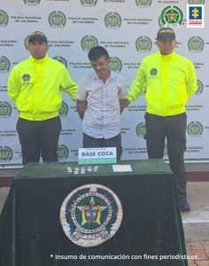 capturado esposado manos atrás custodiado por dos policías. Delante mesa con material incautado. Detrás banner de la Policía Nacional.