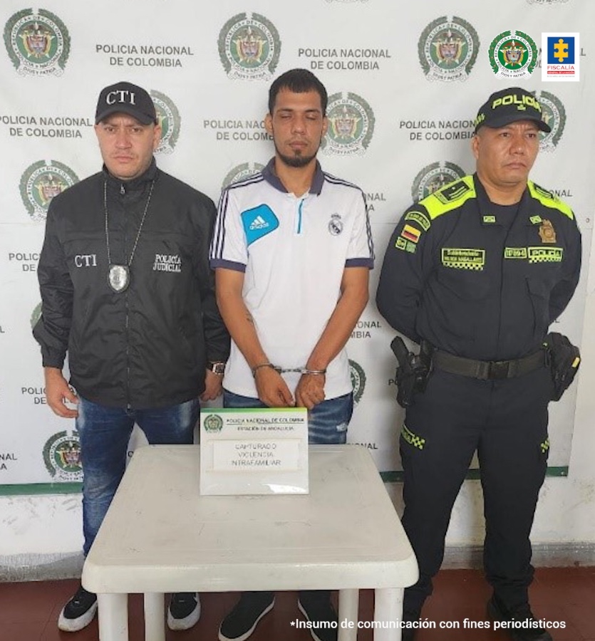 En la fotografía aparece de pie y con los brazos adelante y esposado Sebastián Holguín Guzmán. Presenta barba Viste Camisa de manga corta blanca con rayas azules en los hombros y en los bordes de las mangas; jean azul. Al lado derecho de él hay un servidor del CTI de la Fiscalía. A la derecha de él hay un uniformado de la Policía Nacional. Delante de ellos hay una mesa blanca cuadrada y sobre ella un letrero que dice: ‘Capturado violencia intrafamiliar’. Detrás de ellos hay un pendón institucional de la Policía.