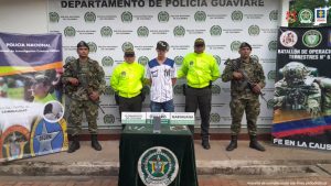 capturado de frente con gorra manos atrás esposado, custodiado por uniformados de la Policía y del Ejército Nacional, delante mesa con material incautado, detrás banner de la Policía y del Ejército Nacional.
