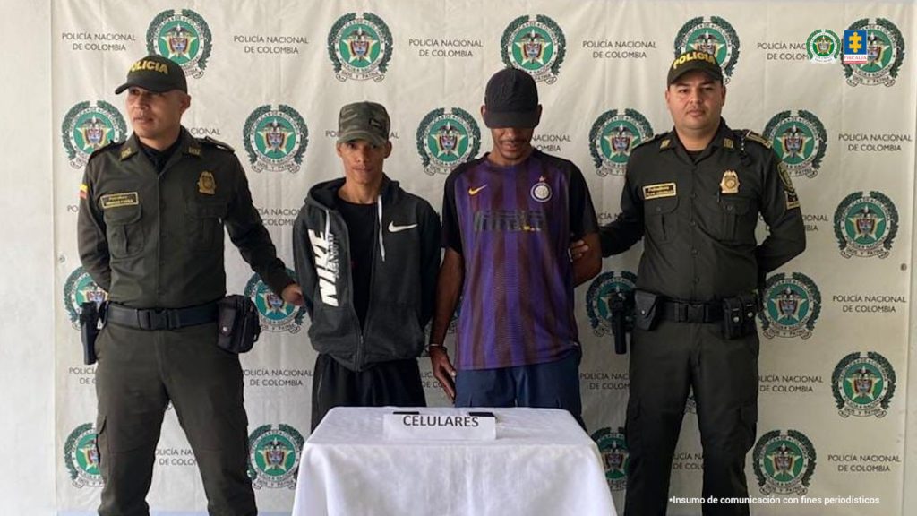 En la fotografía aparecen de pie y con los brazos atrás Sebastián Rengifo Cuellar y Daniel Alfonso Pizarro Rengifo. Uno, usa gorra de color verde; viste con chaqueta de color negro, con estampado blanco en el lado derecho; pantalón negro y zapatillas negras. El otro, usa gorra de color negro; viste con camisa de color púrpura, pantalón azul y zapatillas negras. A los extremos de ellos hay uniformados de la Policía Nacional. Delante de ellos hay una mesa con mantel blanco y sobre ella un letrero que dice: ‘Celulares’. Detrás de ellos hay un pendón institucional de la Policía.