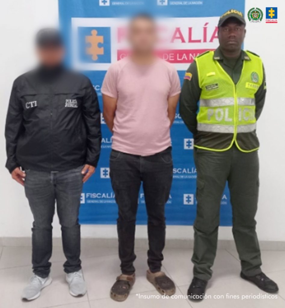 Se observa al capturado de frente, con el rostro bajo un efecto de difuminado. A cada costado un funcionario del Cuerpo Técnico de Investigación CTI y al otro uno de Policía Nacional. Atrás un pendón de la Fiscalía General de la Nación