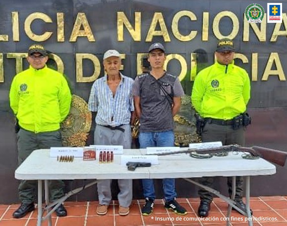 En la fotografía aparecen dos capturados, junto a personal de Policía Nacional y Fiscalía General de la Nación. En la parte posterior de la imagen se ven banners de Policía Nacional y la Fiscalía General de la Nación.