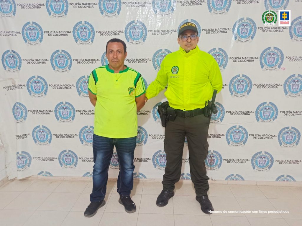 En la fotografía aparece un hombre capturado, acompañado de un uniformado de la Policía. En la parte posterior un banner con logos de la entidad.