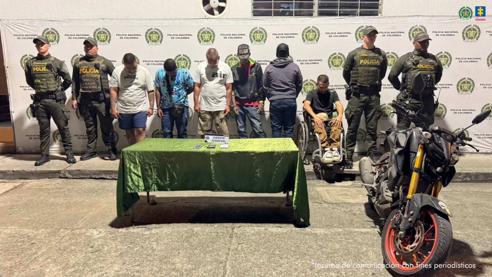 En la fotografía aparecen en hilera los siete judicializados, uno de espaldas a la cámara y otro en silla de ruedas. A los extremos de ellos hay uniformados de la Policía Nacional. Delante de ellos hay una mesa con mantel de color verde oscuro y sobre ella hay un letrero que dice: ‘Orden judicial’ y tres teléfonos celulares. Detrás de ellos hay un pendón institucional de la Policía.