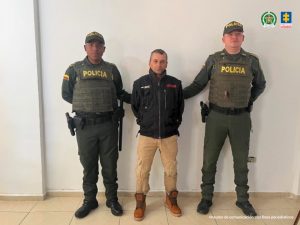 En la fotografía aparece de pie y con los brazos atrás Silvio De Jesús Hurtado Castaño. Viste con chaqueta de color negro y estampados rojo y blanco en lado izquierdo y derecho del pechos, respectivamente; pantalón caqui con bolsillos en las rodillas y botas color marró. En sus extremos hay uniformados de la Policía Nacional.