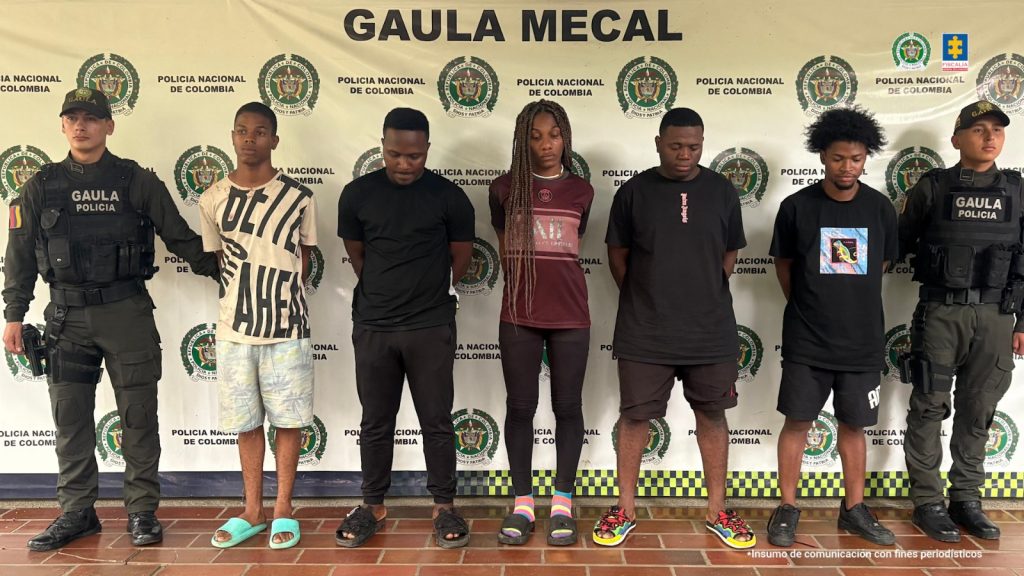 En la fotografía se aprecian de pie y con los brazos atrás a cinco presuntos implicados del secuestro extorsivo de dos hermanos. A los extremos de ellos hay uniformados de la Policía Nacional y detrás de ellos hay un pendón institucional del Gaula de la Policía.