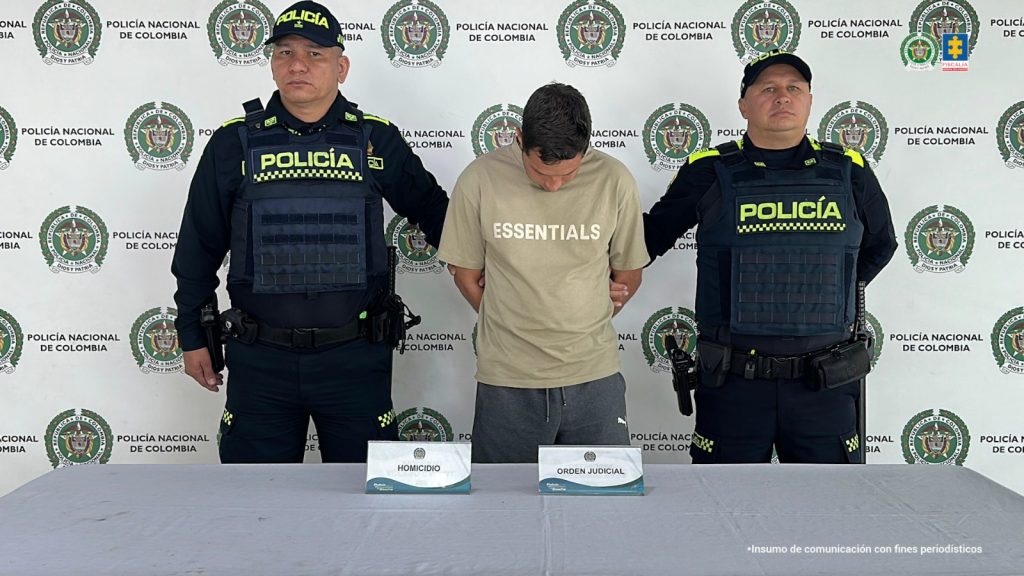 Fotografia en donde se puede ver al capturado en medio de dos policias, detras de ellos se encuentra un pendon de la policia y delante de ellos se ven los elementos incautados sobre una mesa