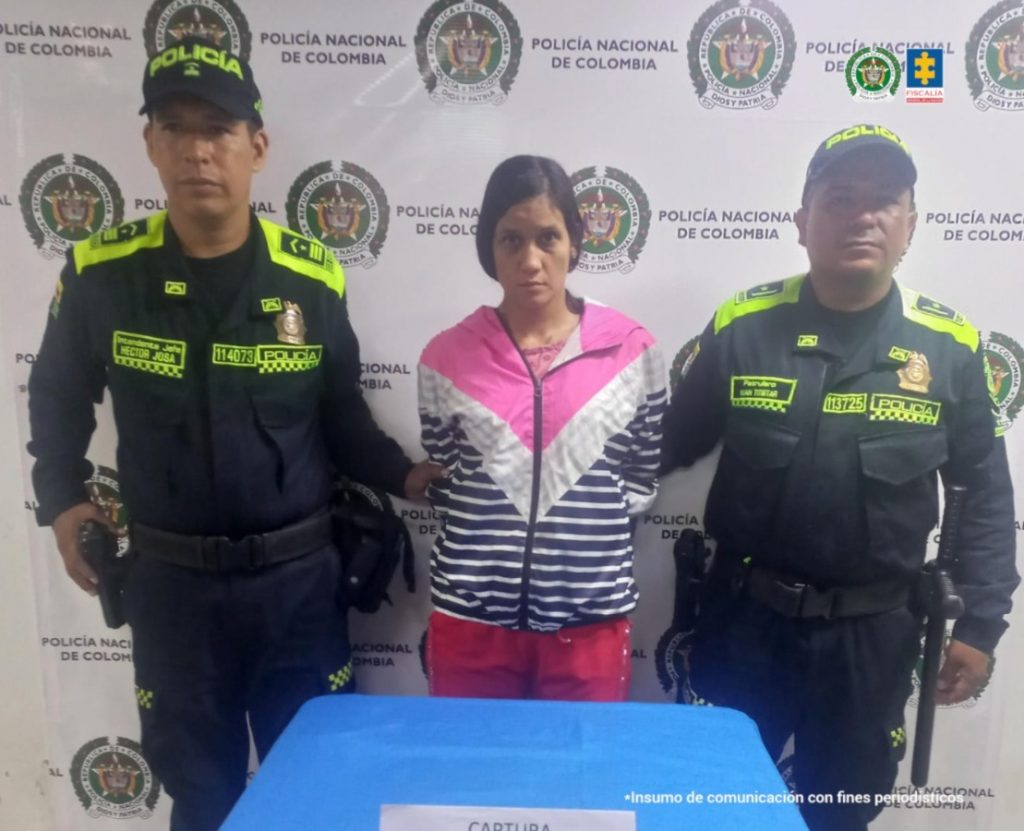 En la fotografía se aprecia de pie y con los brazos atrás a Stephani Cardona Pedroza. Viste con buso de sudadera con franjas anchas y delgadas de colores fucsia, blanco y azul oscuro; así como pantalón rojo. A sus extremos hay uniformados de la Policía Nacional. Detrás de ellos hay un pendón institucional de la Policía.