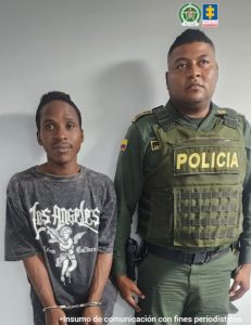 En la fotografía se aprecia de pie, con los brazos adelante y esposado a Stiven Alberto Vallecilla. Viste con camiseta gris de manga corta; bermuda gris desgastada y pantuflas de color gris. A su izquierda hay un uniformado de la Policía Nacional.