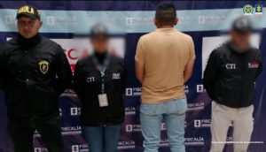 En la fotografía aparece el capturado a dos servidores del CTI y un agente del Gaula Militar. En la parte superior está un banner de la Fiscalía General