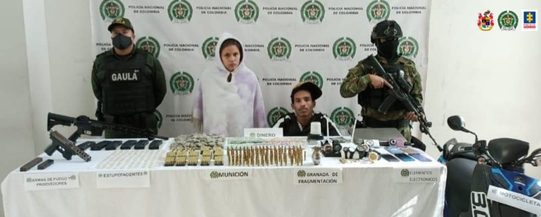 En la fotografía se observan a los 2 capturados junto a dos agentes de la Policía Nacional. En la parte posterior se observa una mesa con armas de fuego, proveedores, munición, explosivos, teléfonos celulares y una motocicleta