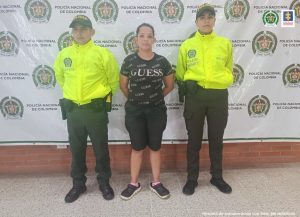 capturada en bermuda de frente, manos atrás esposadas, custodiada por dos uniformados de la Policía. Detrás banner de la Policía Nacional.