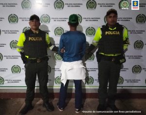 capturado de espalda custodiado por dos policías, detrás banner de la Policía Nacional.
