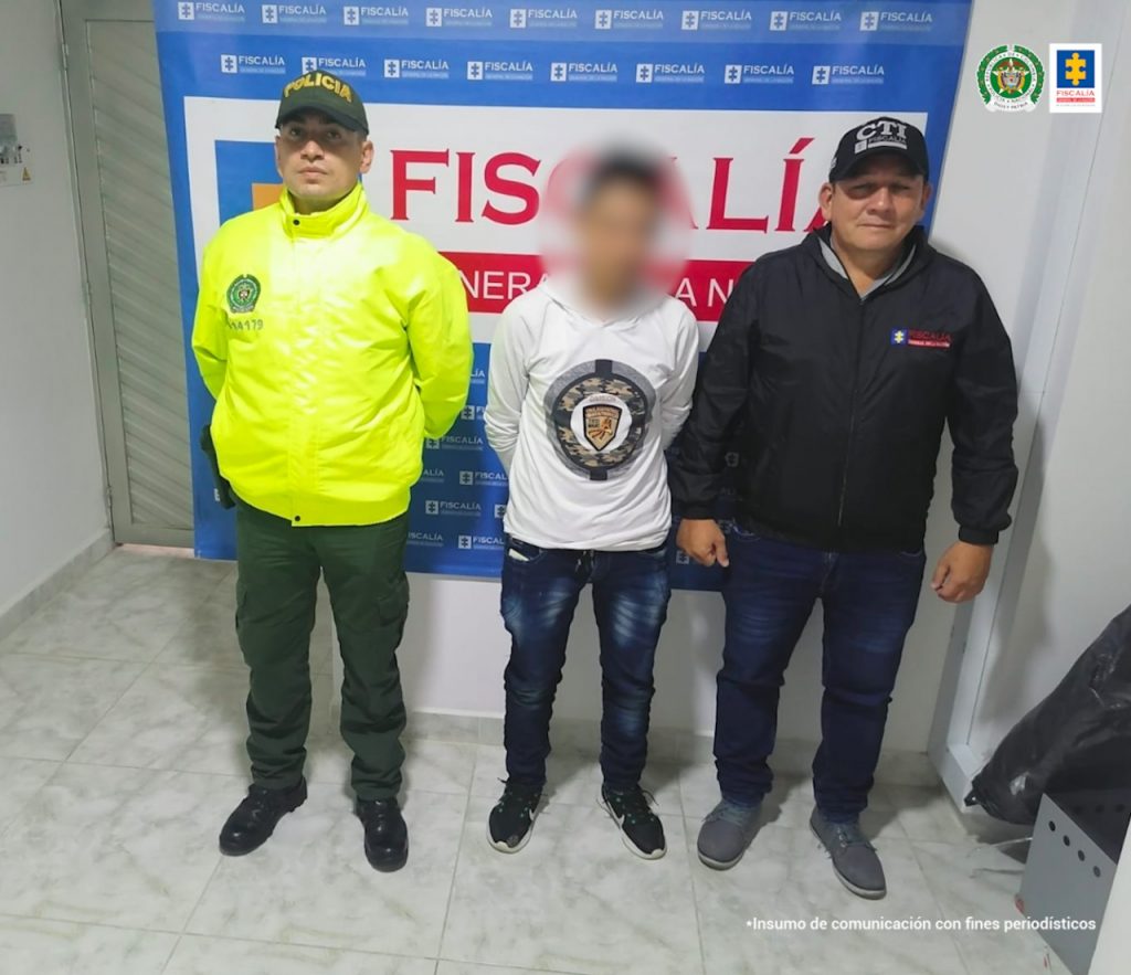 En las fotografía aparece un capturado junto a personal de Fiscalía General de La Nación y Policía Nacional. En la parte posterior de la imagen se ven banners de Policía Nacional y la Fiscalía General de la Nación.