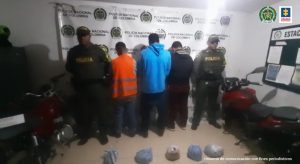 En la foto aparecen de pie y de espaldas los tres presuntos implicados en transportar 10 kilos de marihuana. A lado y lado de ellos hay uniformados de la Policía Nacional y se aprecian dos motocicletas. En la parte posterior hay un pendón institucional de la Policía Nacional.