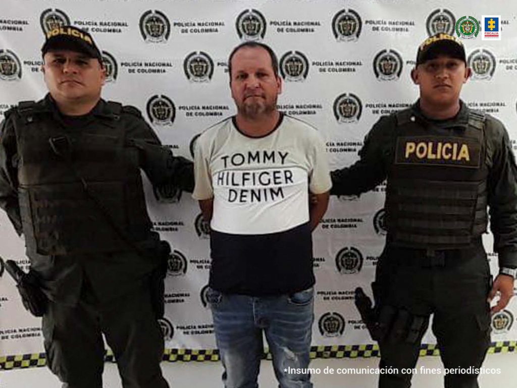 capturado de frente manos atrás esposado, custodiado por dos policías. Detrás banner de la Policía Nacional.