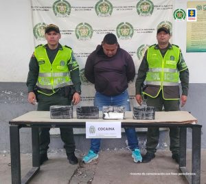 Dos uniformados de la Policía Nacional custodian al procesado, de camisa negra, quien está esposado. En la mesa está el estupefaciente que fue incautado.Atentamente,