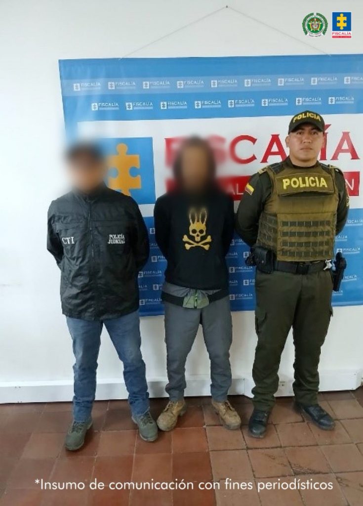 En la fotografía aparece de pie y con los brazos atrás el hoy imputado. Usa cabello largo, y bigote poblado. Viste con buso negro, con estampado amarillo en el pecho; pantalón de color gris y zapatos marrón. A su extremo derecho hay un servidor del CTI de la Fiscalía y a la derecha un uniformado de la Policía Nacional. Detrás de ellos hay un pendón institucional de la Fiscalía.