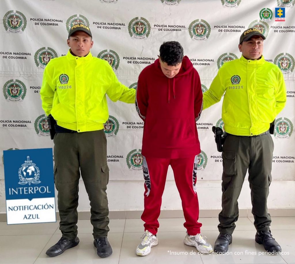 Se observa al capturado de frente con la cabeza abajo, viste chaqueta y pantalón rojo, zapatos tenis blancos. A cada lado lo custodia un uniformado de la Policía Nacional. Atrás, un pendón de Policía Nacional y sobrepuesto un texto que dice INTERPOL NOTIFICACIÓN AZUL.