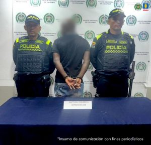 Se observa al capturado de espaldas y un efecto difumina su cabeza. A cada lado lo custodia un uniformado de Policía Nacional. Al frente, sobre una mesa está un aviso que dice Captura Violencia Intrafamiliar.