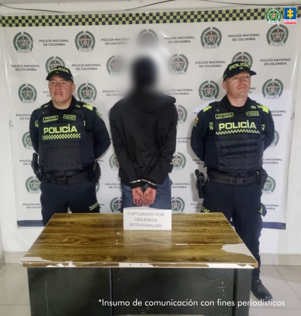 Se observa al capturado de espaldas y a cada costado un funcionario de Policía Nacional custodiando. Al frente una mesa que dice VIOLENCIA INTRAFAMILIAR, atrás un pendón de Policía Nacional.