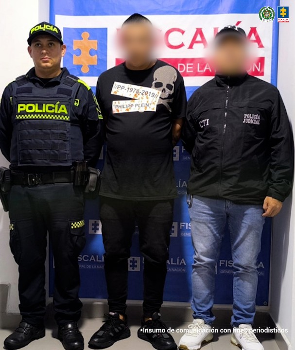 Se observa a un hombre capturado, de frente y sobre su rostro un efecto que lo hace difuminado. Lo custodian un uniformado del CTI y otro de Policía nacional. Atrás, un pendón de la Fiscalía General de la Nación