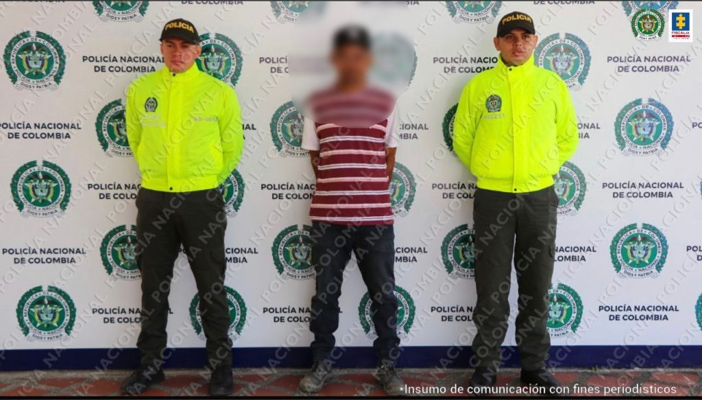 Se observa el capturado en el centro y a cada lado un funcionario de Policía Nacional. Atrás un pendón de Policía Nacional