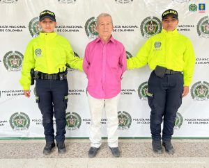 El imputado se encuentra en custodia por un uniformado de la Policía Nacional. El capturado tiene la cara cubierta y tiene un buzo blanco. Detrás se observa un backing de la Policía.