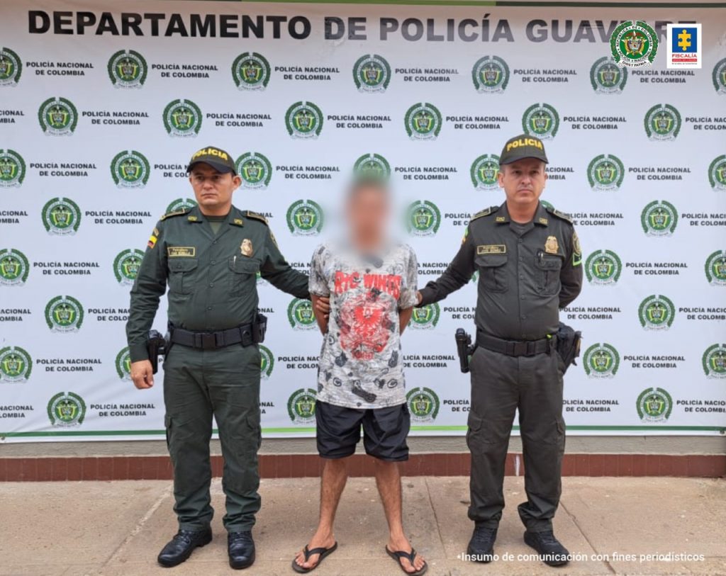 capturado de frente en pantaloneta y chancletas custodiado por dos uniformados de la Policía. Detrás banner de la Policía.