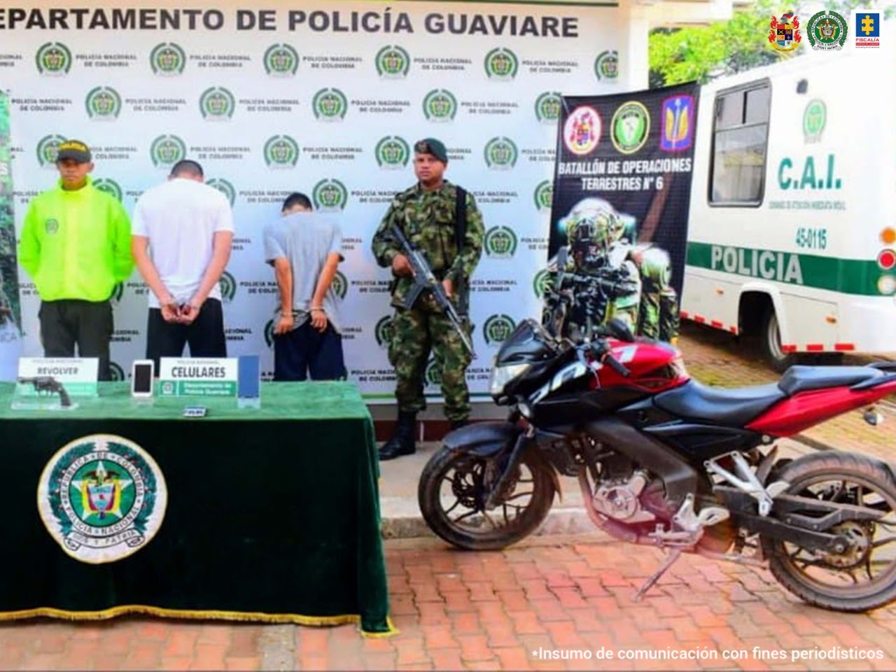 en la foto aparecen de espaldas dos hombres capturados, custodiados por un uniformado de la Policía y por un militar del Ejército Nacional detrás y al lado de estas personas hay un banner de la Policía, otro del Ejército y un CAI móvil. Delante de ellos hay una mesa con los elementos incautados en la diligencia y la motocicleta en la que se movilizaban los detenidos.
