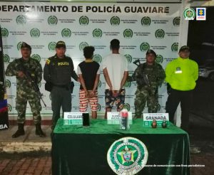 capturados de espalda, custodiados por uniformados de la Policía y del Ejército Nacional. Detrás banner de la Policía Nacional, delante de ellos una mesa con el material incautado.