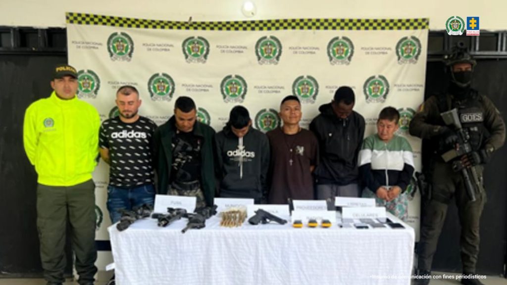 En la fotografía se aprecian de pie y en hilera a seis hombres. A sus extremos hay uniformados de la Policía Nacional. Delante de ellos hay una mesa con mantel blanco y sobre ella hay armas de fuego cortas y largas, munición, radios de comunicaciones y celulares. En el suelo hay prendas militares. Detrás de ellos hay un pendón institucional de la Policía.