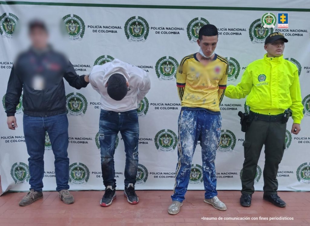 En la foto se observa a los capturados custodiados por un servidor del Cuerpo Técnico de Investigación CTI y un uniformado de la Policía Nacional. También se observa un backing de la Policía Nacional. Uno de los capturados tiene la cabeza agachada y el otro tiene una sudadera manchada con pintura y una camiseta de la selección Colombia.