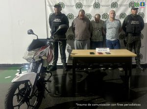 En la imagen están los 3 procesados con dos servidores del Gaula de la Policía Nacional, una mesa con 3 celulares y al lado la motocicleta incautada. 