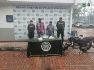 capturados de frente esposados, custodiados por dos uniformados de la Policía, delante de ellos una mesa con droga y al lado motocicleta incautados, detrás banner de la Policía Nacional.