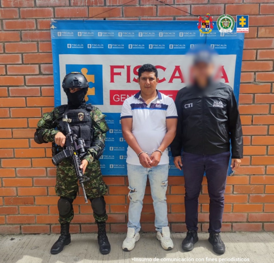 El capturado se encuentra de frente y custodiado por un servidor del Cuerpo Técnico de Investigación CTI y un uniformado del Ejército Nacional. Detrás de la foto hay un backing de la Fiscalía General de La Nación.