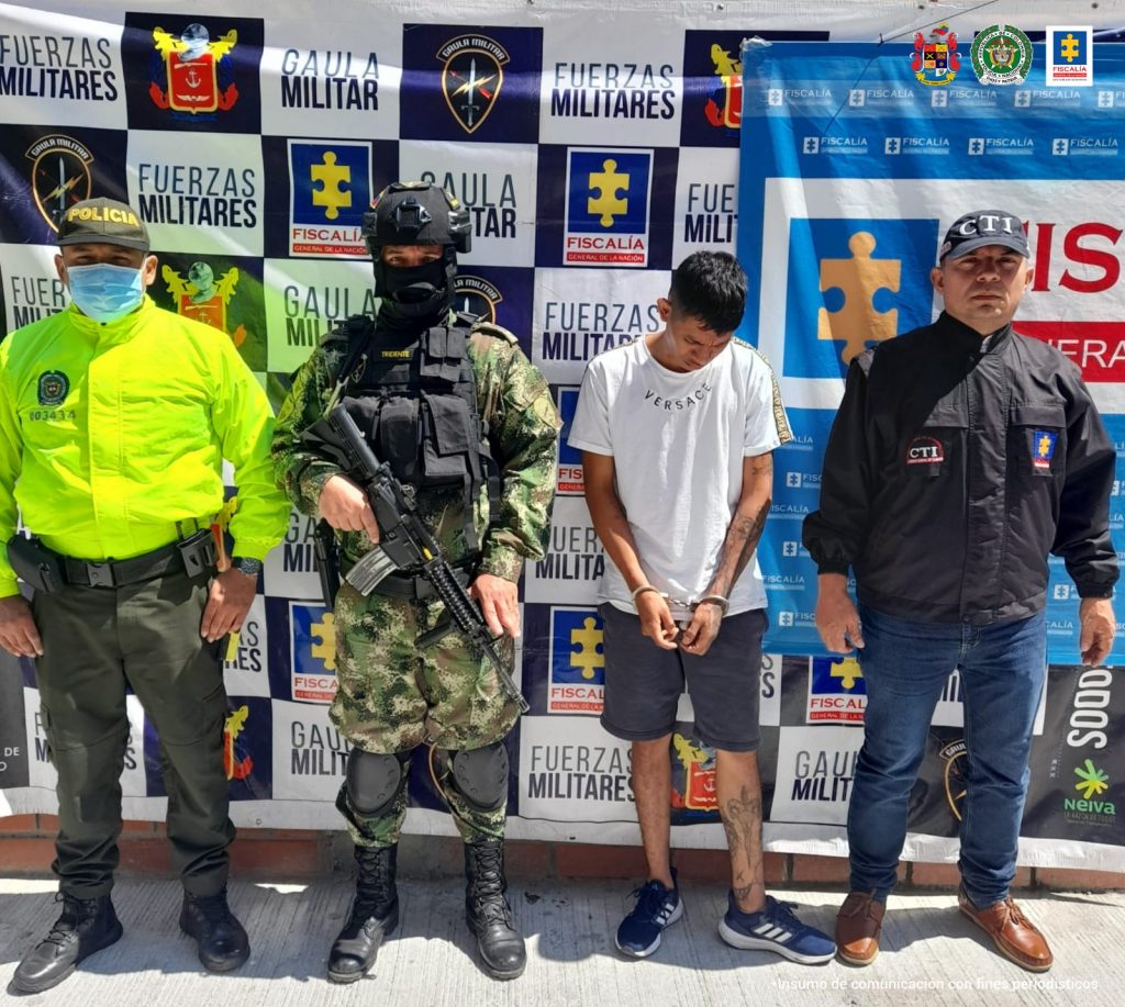 En la fotografía aparece un capturado, junto a personal de Fiscalía General de la Nación, Ejército Nacional y Policía Nacional. En la parte posterior de la imagen se ven banners de Ejército Nacional, Policía Nacional y la Fiscalía General de la Nación.